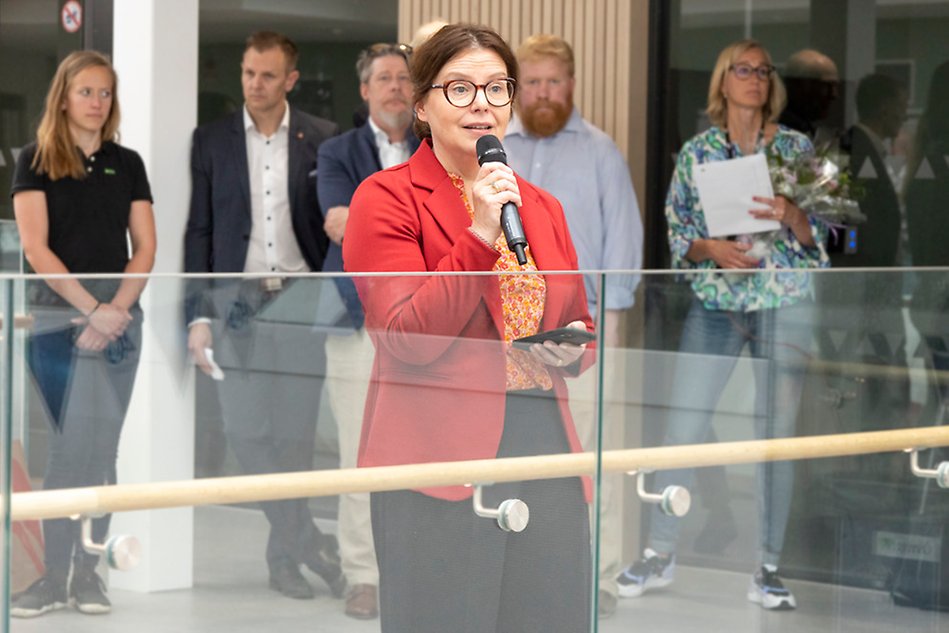 Woman in brown hair wears a red blazer and glasses and speaks in a microphone. There are people in the background who are listening. Photo.
