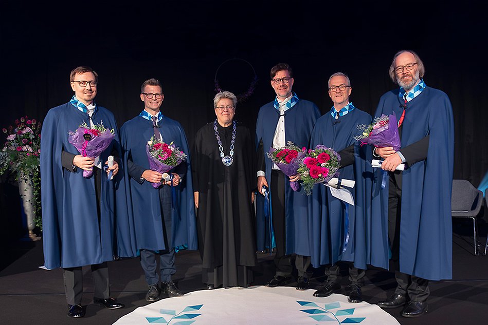 Fem personer i blå caper står uppradade på en scen. I famnen har de blommor och diplom. I mitten står en kvinna i svart rektorskappa och med en stor silverkedja runt halsen Foto.