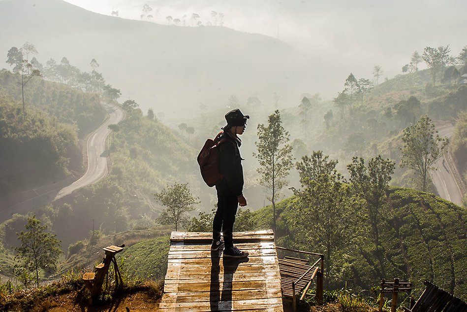 En person står på en bro framför ett vidsträckt landskap av dimmiga berg, träd och en grusväg. Foto.