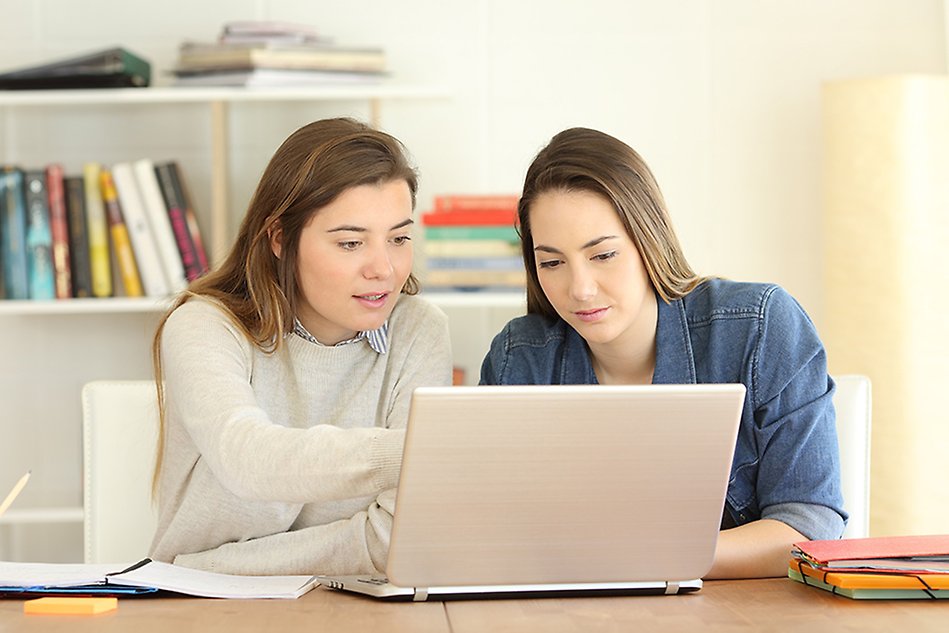 Två unga kvinnor sitter vid ett skrivbord framför en laptop. Den ena kvinna pekar på något på skärmen. Foto.
