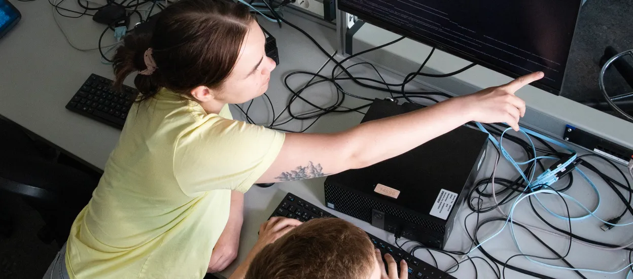 A young girl is leaning over a desk full of cables, pointing at a screen. Photo.