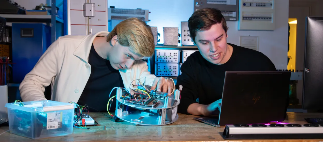 Two people by a table, one is poking at a steel structure with cables lying on the table and the other is typing on a computer. Photo.