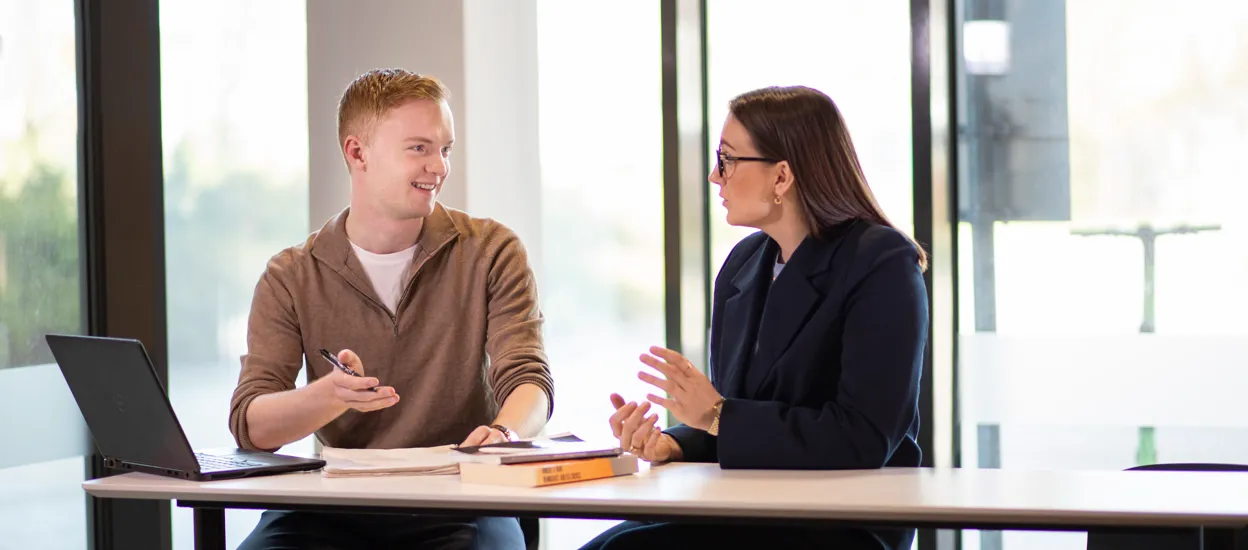 Two people are talking to each other at a table with a computer and books in front of them. Photo.