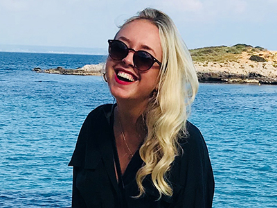 Porträtt av en ung, leende kvinna med solglasögon och svart klänning. I bakgrunden syns en ö i ett grönblått hav. Foto.