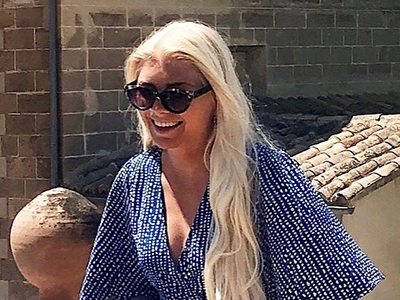 En blond ung kvinna med långt hår och solglasögon står framför en brun tegelvägg. Foto.
