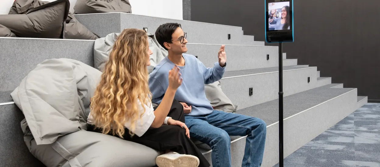 Two young people are sitting at a classroom platform, waving at a person in a videocall. Photo.