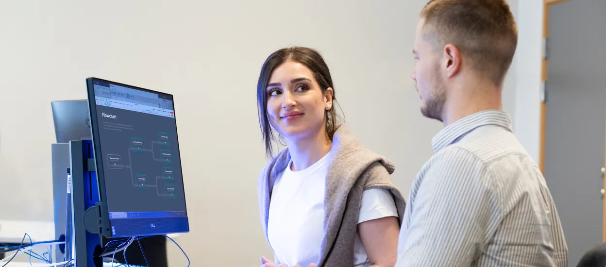 Two young people are interacting by a desk with a computer screen in front of them. Photo.