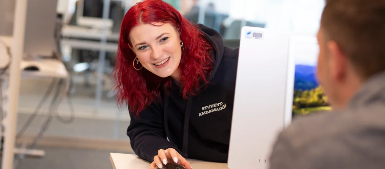 A young woman looks out from behind a computer screen at a blurred person in the foreground. Photo.