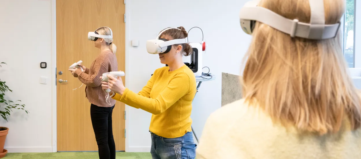 Three women wearing white VR goggles stand in a bright room. Photo.