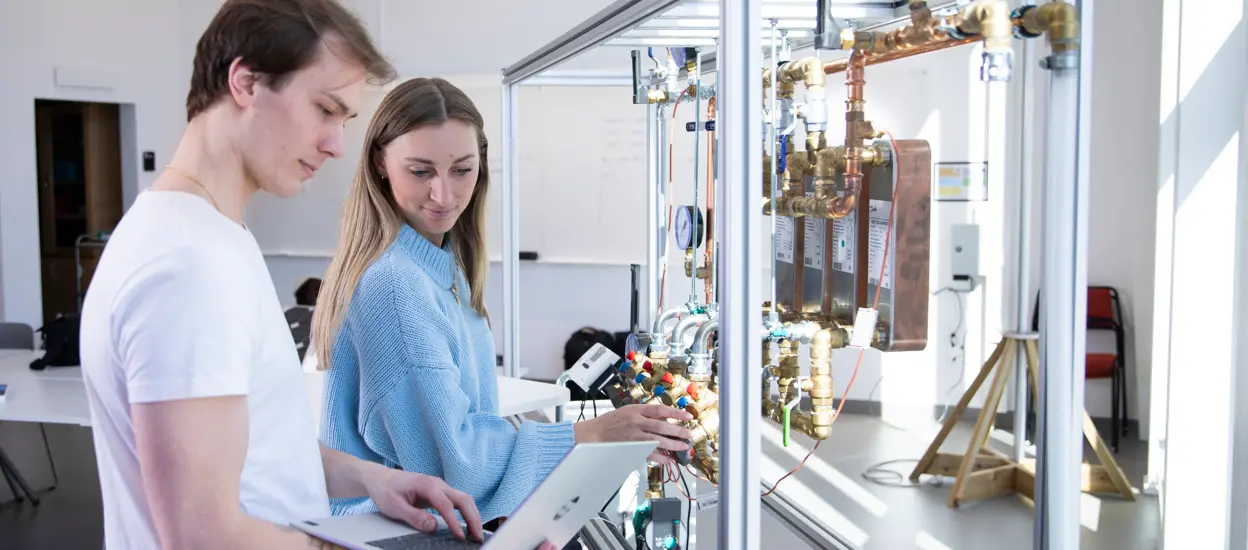 Two young students are inspecting taps in a lab environment. One of them is typing on a laptop. Photo.