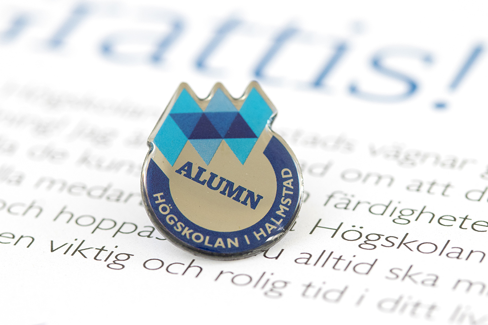 Close-up of a pin with the Halmstad University logo and the word "alumn". Photo.