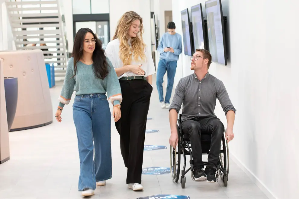 Tre unga personer i en korridor. En person sitter i rullstol. I bakgrunden syns ytterligare en person. Foto