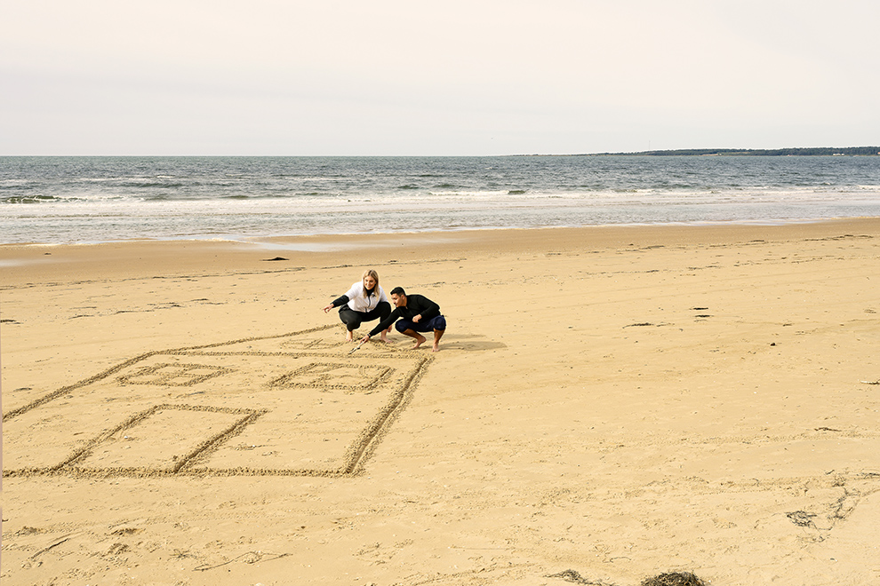 Två personer ritar ett stort hus i sanden på en strand. I bakgrunden syns havet. Foto. 
