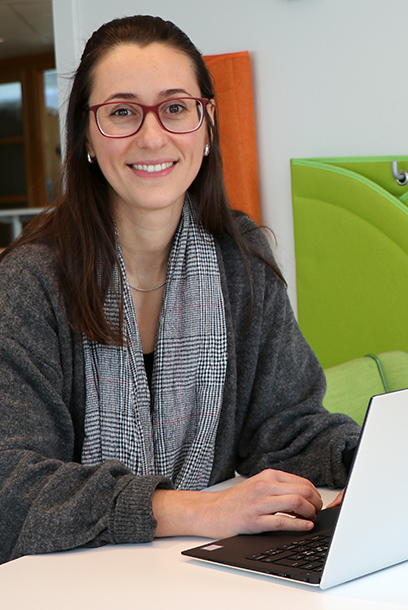 Smiling woman with dark hair and glasses sitting at a table with a laptop in front of her. Photo.
