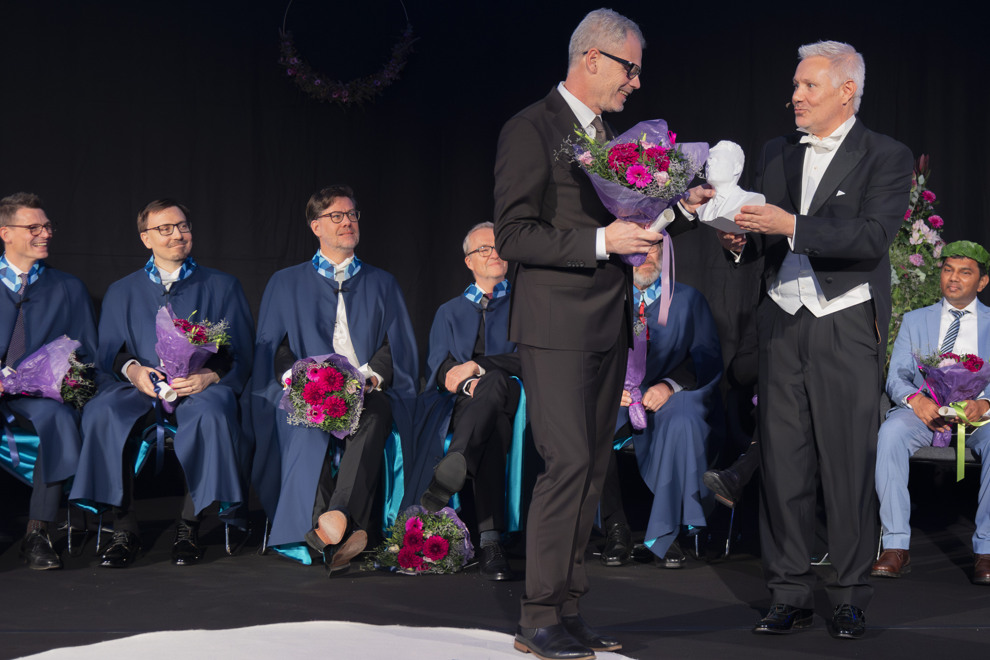 Två finklädda personer på en scen. Den ena ger den andra ett pris och en bukett blommor. Flera personer sitter i bakgrunden. Foto.