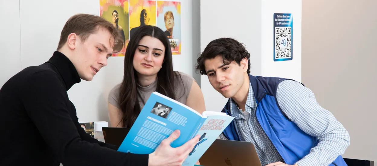 Tre studenter sitter vid ett bord och tittar i en bok. Foto.