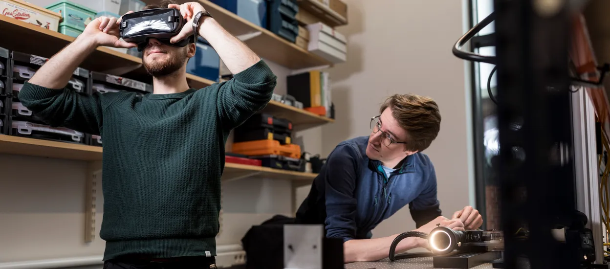 Två studenter i ett labb med lådor, pärmar och sladdar. Ena studenten har VR-glasögon på sig och den andra tittar på honom. Foto.