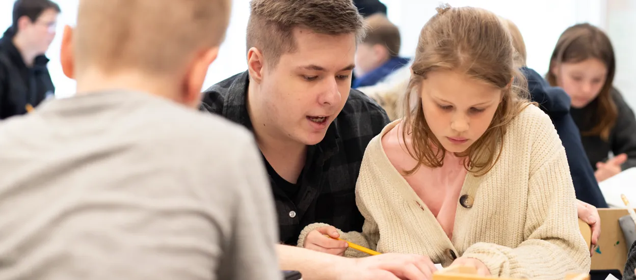 En manlig lärarstudent sitter på huk vid en flicka och han hjälper henne i klassrumsmiljö.