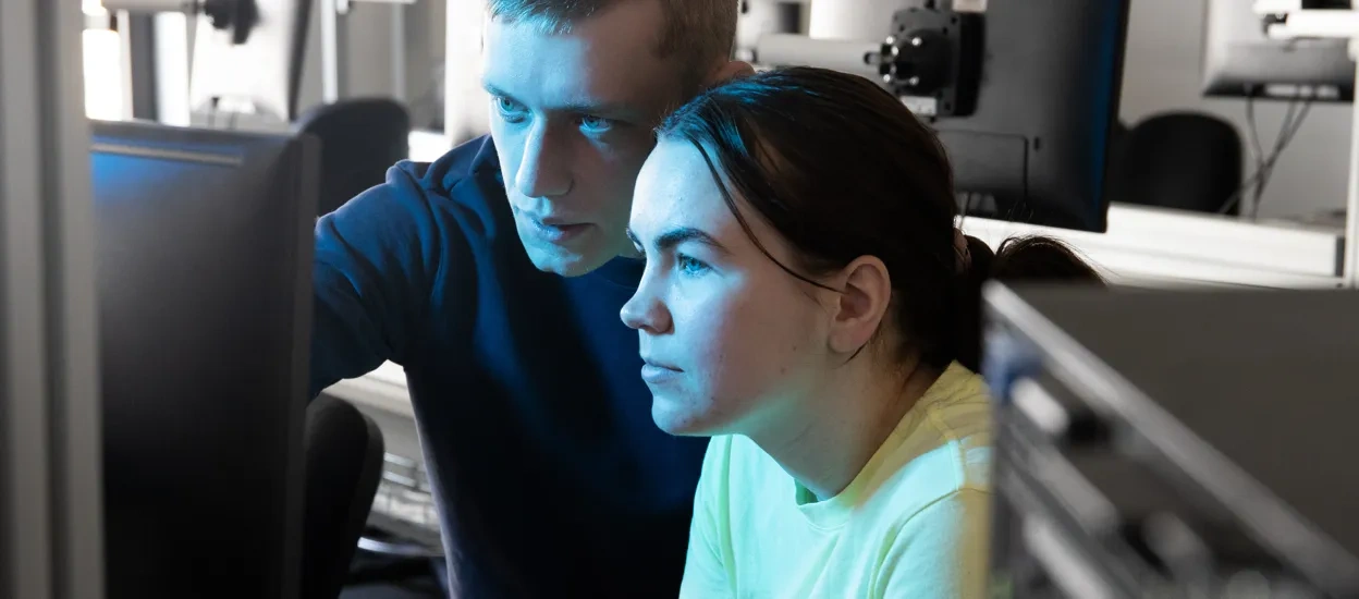 Två studenter sitter vid en skärm i en datorsal. Det blåa ljuset från skärmen reflekteras i deras ansikten. Foto.