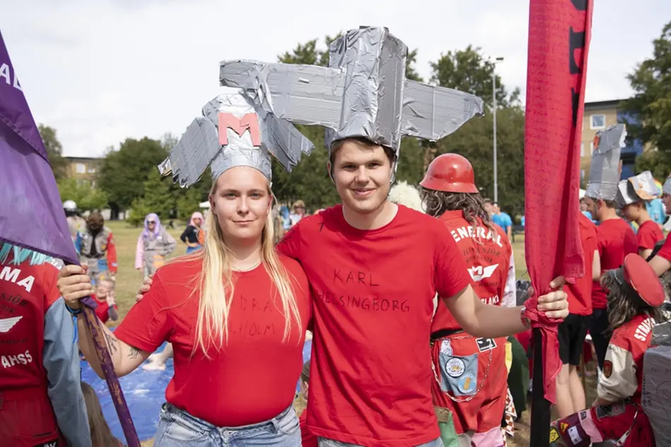 En ung kvinna och en ung man står bredvid varandra och ler. De har röda t-shirtar och håller flaggor i sina händer. Foto.