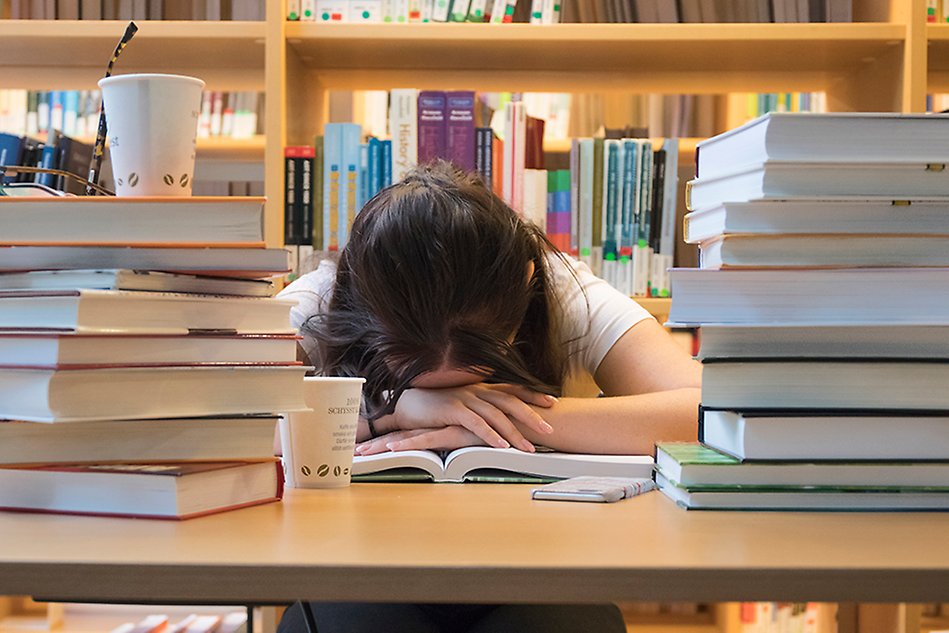 A person is sitting at a table, surrounded by book stacks, with her face down in a book. Photo.