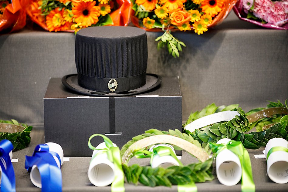 Table with doctoral hat, wreaths and diplomas wrapped with coloured ribbons. Photo.