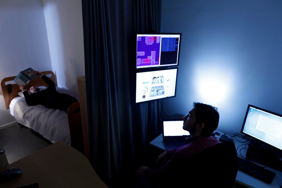 En man ligger och läser en tidning i en säng, en annan man övervakar med hjälp av datorskärmar. Foto.