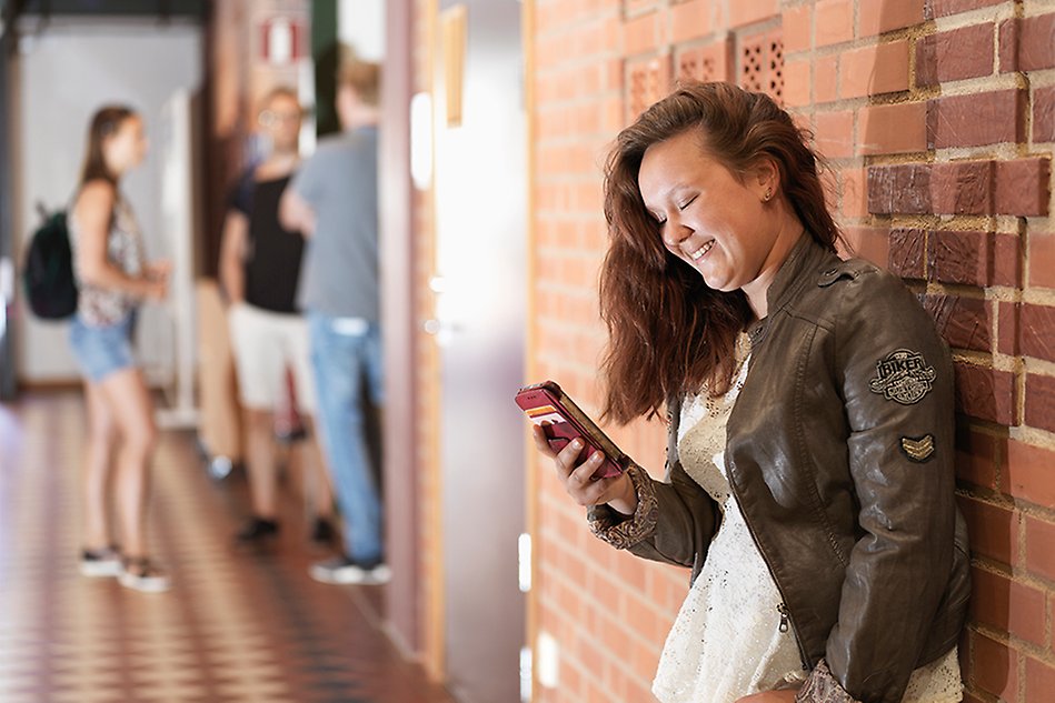 Ung, kvinnlig student står lutad mot en röd tegelvägg och tittar ned i sin mobiltelefon. I bakgrunden står en grupp andra, glada studenter. Foto. 
