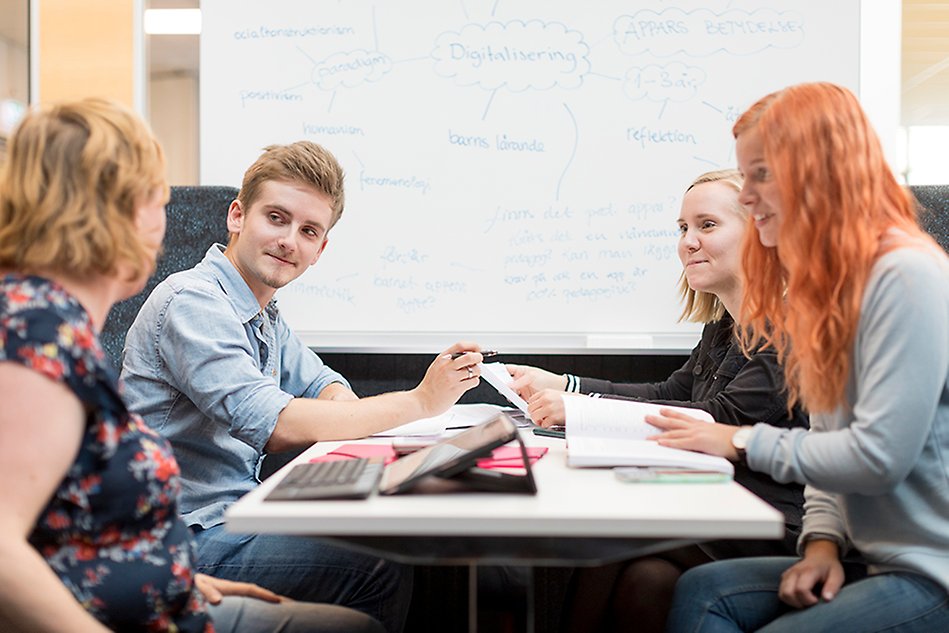 Four people sit around a table full of books and talk to each other. Three of them are looking at the fourth person who is in the lower left corner. Behind them is a whiteboard with text. Photo.