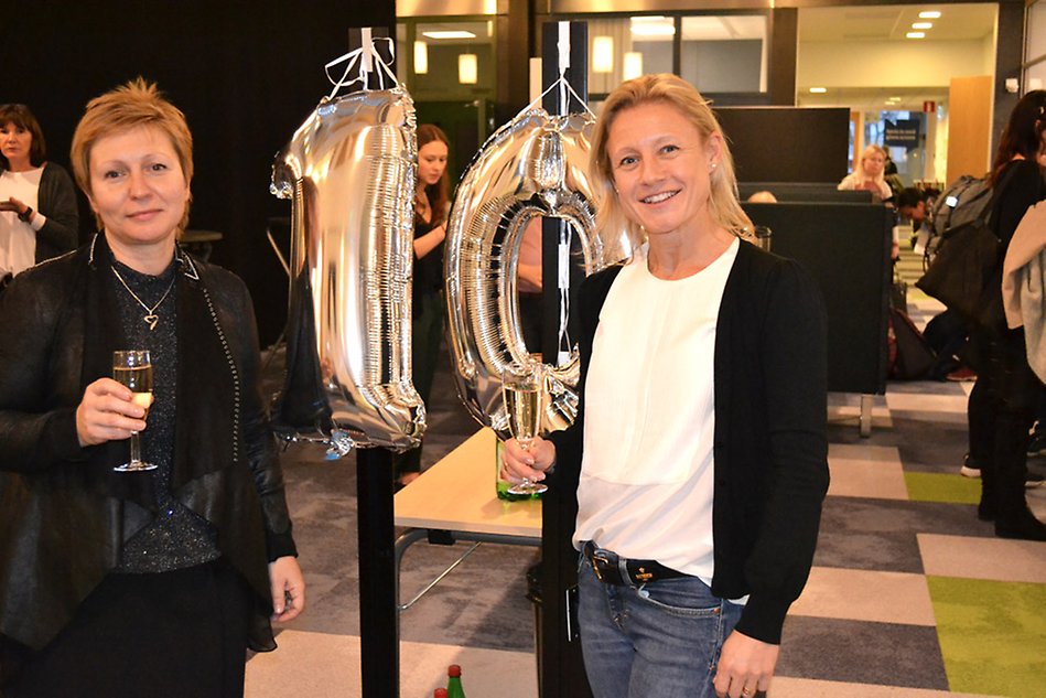 Två kvinnor står med bubbelglas framför silverballonger i form av talet 10. 