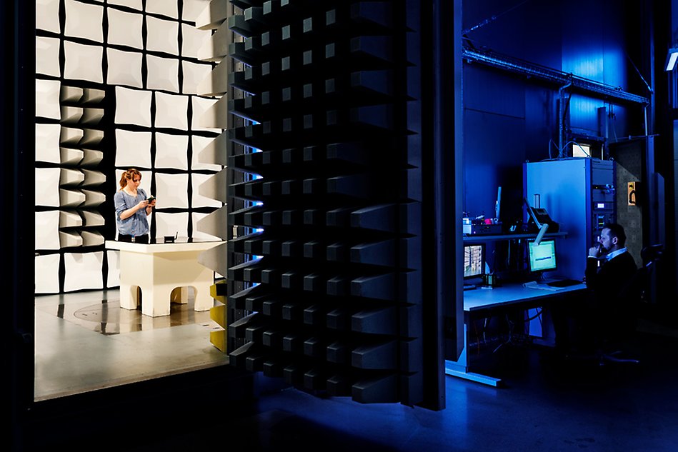 Woman standing at table inside sound-insulating chamber and holding an electronic device while outside sitting a man and studying a computer screen. Photo. 