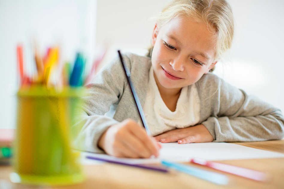 Colourful pens, blonde girl writing at a table. Photo.