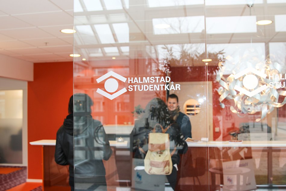 Studentkårens reception där två studenter får hjälp. Bilden är tagen genom en glasvägg så att kårens logotyp syns på glaset. Foto.
