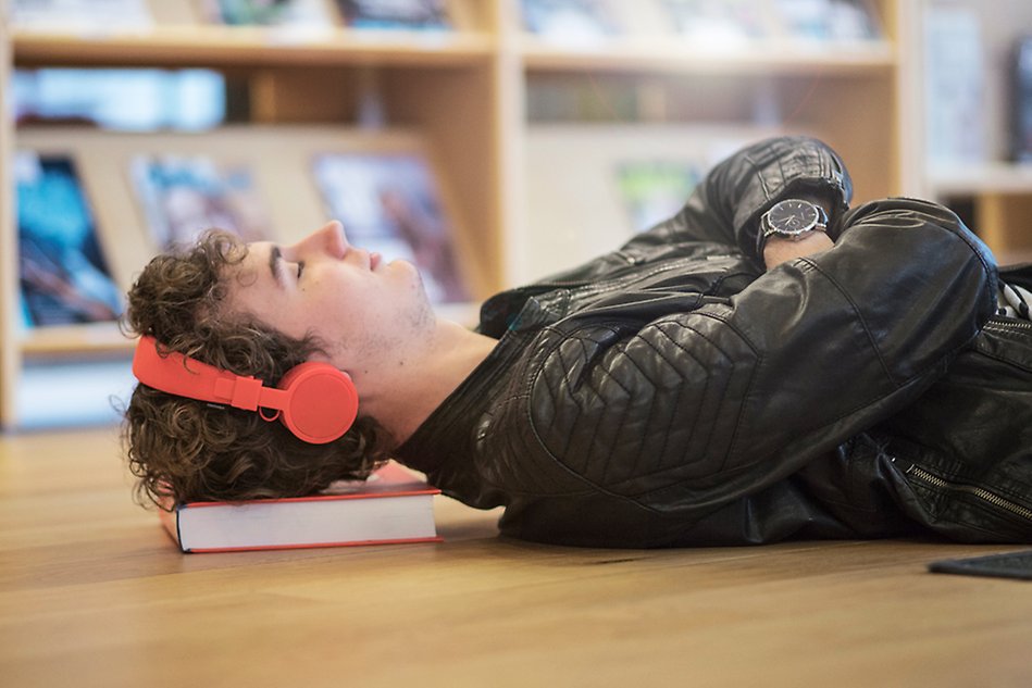 Kille ligger på golvet och blundar, han har röda hörlurar på sig och böcker som kudde. Foto. 