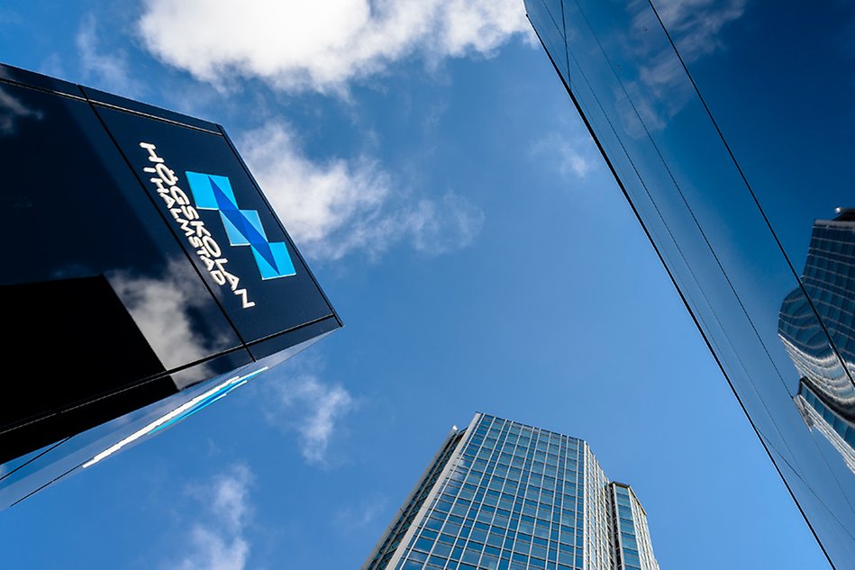 Photo taken from below up against a blue sky with a couple of fog clouds. To the left a black pillar with the University’s logo. In the foreground part of a building in glass and steel, which is reflected in a part of a window to the right.