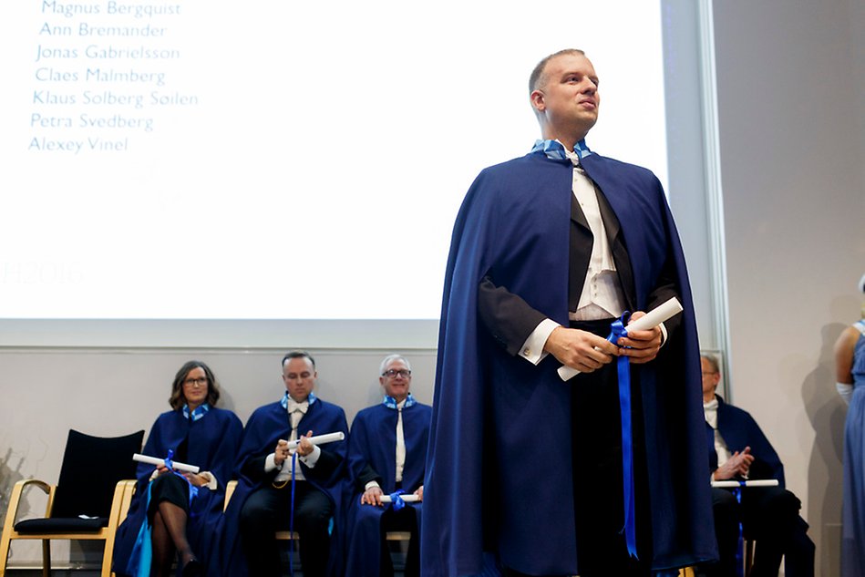 A man wearing a blue cape and holding a diploma is standing in front of a screen projection. A group of other people in capes are sitting behind the man. Photo.