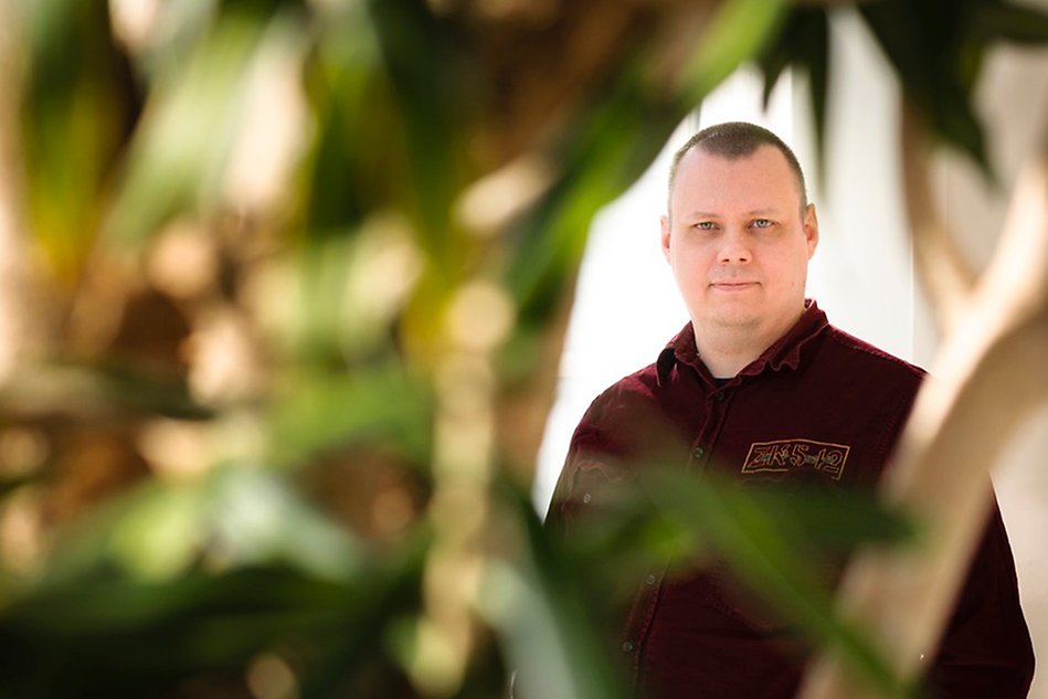 Man with short hair wearing a burgundy shirt, standing next to greenery. Photo.