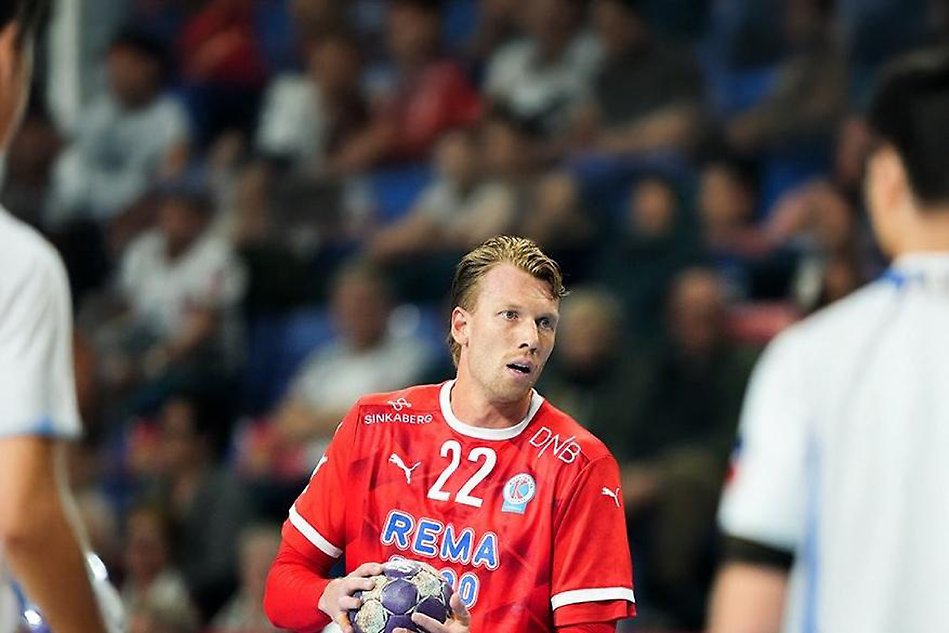 Handbollsspelare i röd tröja full med sponsorer och nummer 22 håller i en handboll. I förgrunden finns motståndare. I bakgrunden finns publik. Foto.