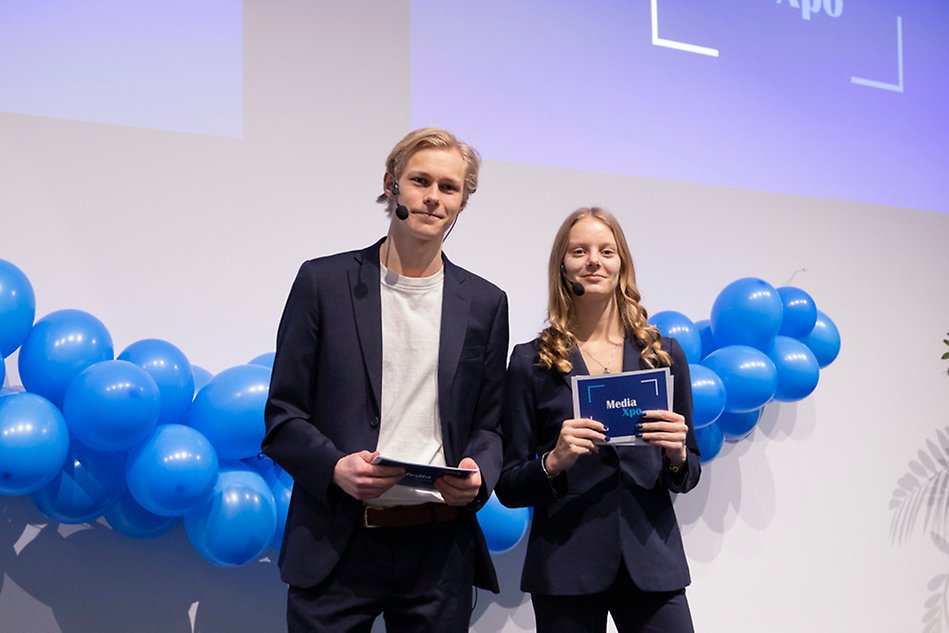 Två studenter i mörk kostym håller i läskort och ler mot kameran. I bakgrunden finns ballonger. Foto.