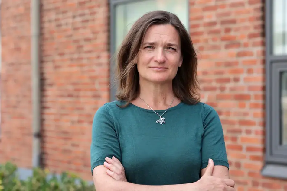 A woman wearing a green top standing in front of a brick wall with her arms folded. Photo.