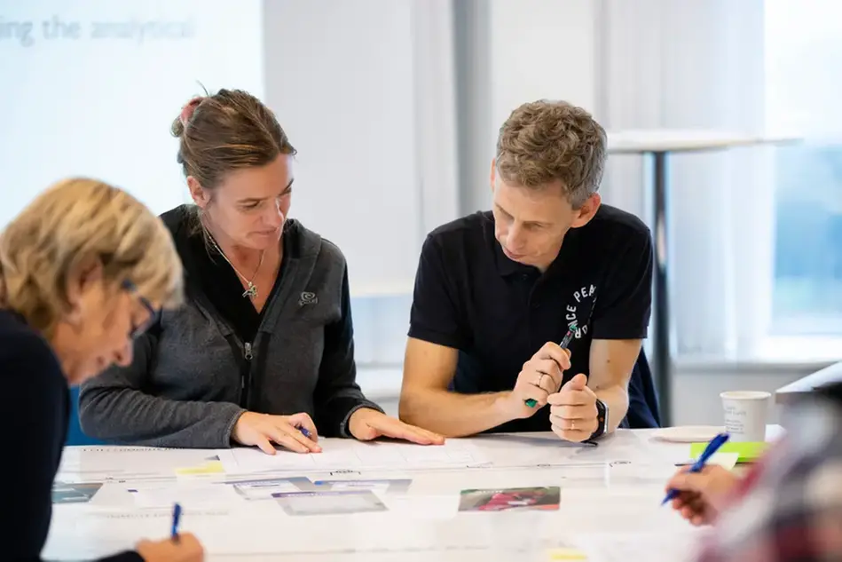 Two people looking down at a table covered with papers and pens. Photo.
