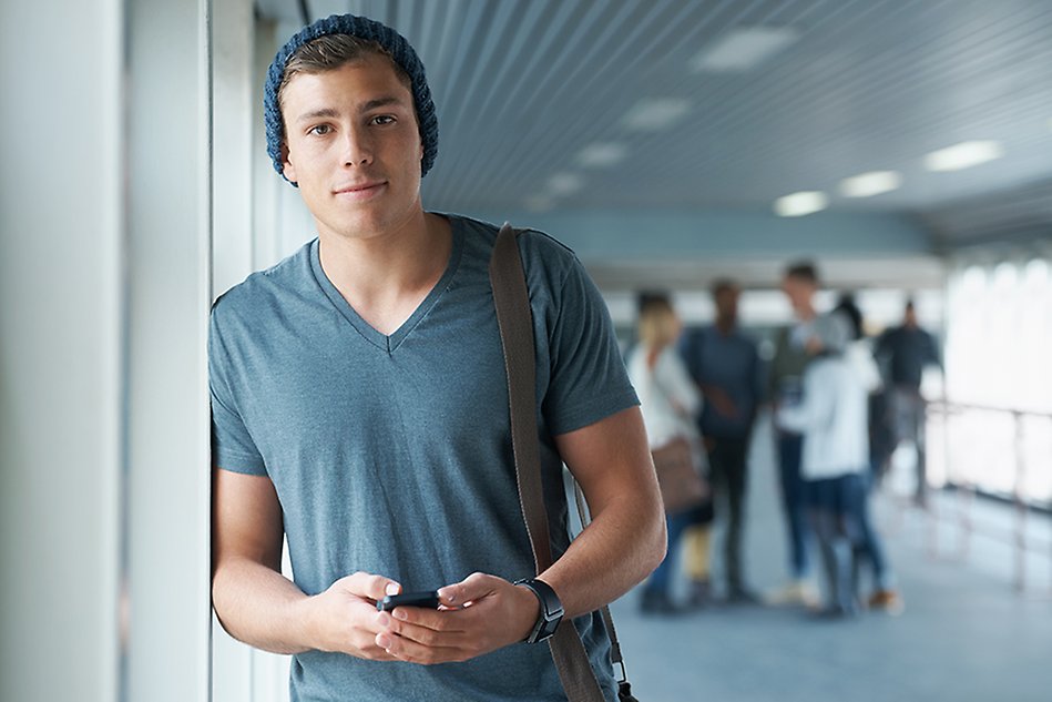 En ung man står lutad mot en vit vägg, håller i en mobiltelefon och tittar in i kameran. Foto. 