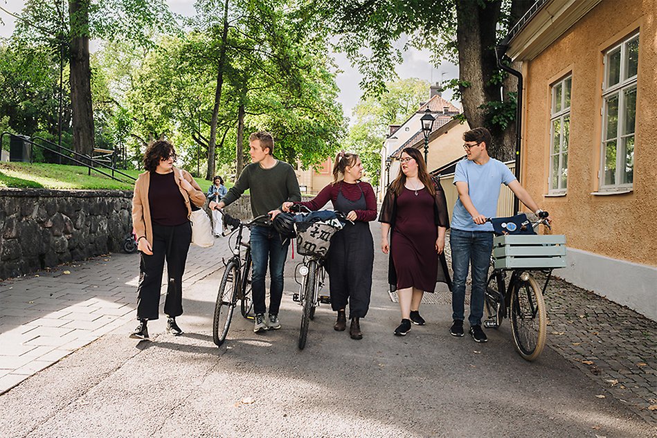 Five people, some with bikes, walking on a small street talking to each other. Photo. 
