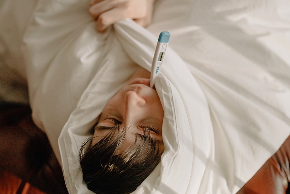 A person is lying down, embedded in a thick blanket and with a fever thermometer in his mouth. Photo.