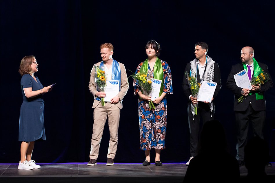 En ung kvinna och tre unga män står på rad på en scen med blommor och diplom i sina händer. De är alla finklädda. Foto.