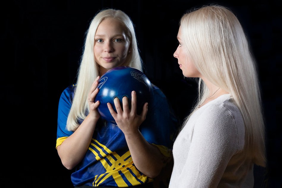 Blond, långhårig tjej i grå tröja tittar på sin spegelbind. Spegelbilden är hon själv som håller i ett bowlingklot och är klädd i blågul landslagströja. Svart bakgrund. Foto.