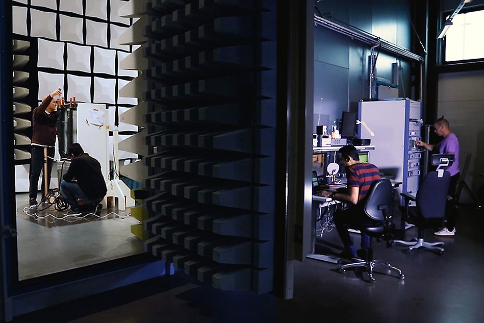 Two people working inside a test chamber for electronic devices, two other people working with equipment outside the chamber. Photo.