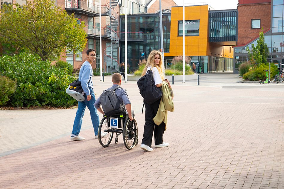 Three people in an outdoor open space, surrounded by bushes and buildings. Photo