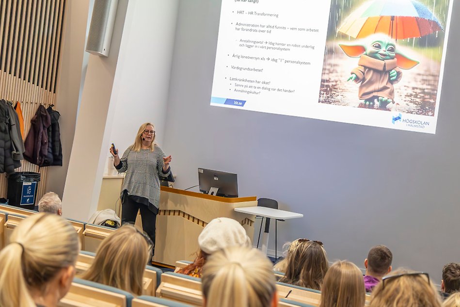 Kvinna i svartvita kläder föreläser inför studenter i en sal. I bakgrunden visas en presentation. Foto.