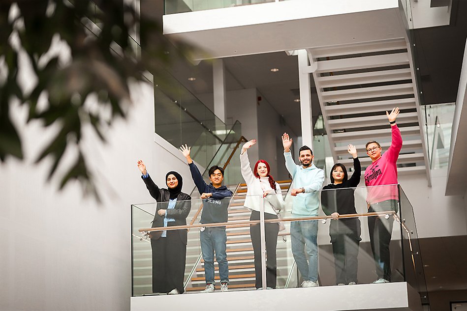 A group of students smiling and waving on a balcony at campus. Photo. 